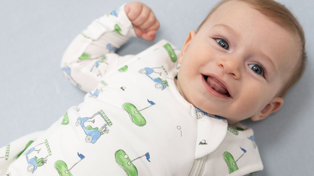 Laughing baby lying on a pale blue sheet in a white zip-up sleep sack printed with golf carts and greens
