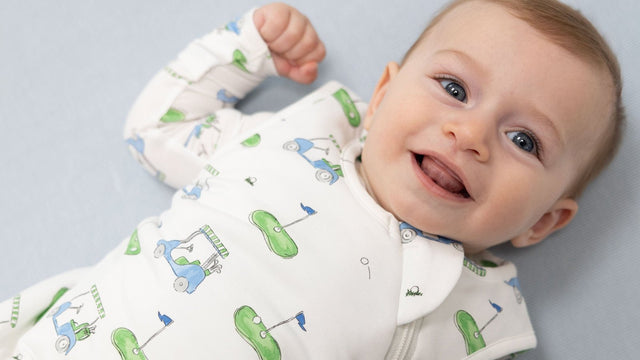 Laughing baby lying on a pale blue sheet in a white zip-up sleep sack printed with golf carts and greens