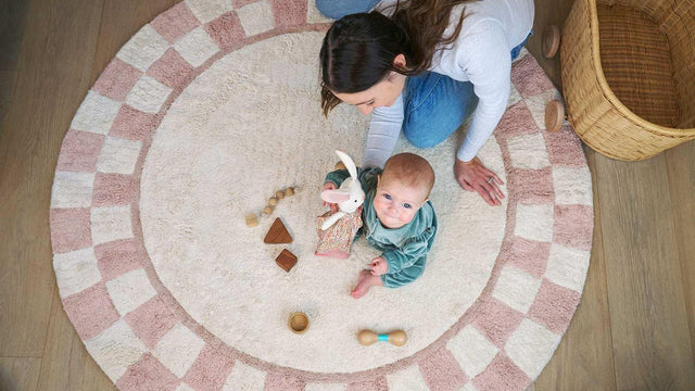 Baby in a green romper sitting on a round pink checkered washable cotton rug with wooden toys, her mother kneeling beside her