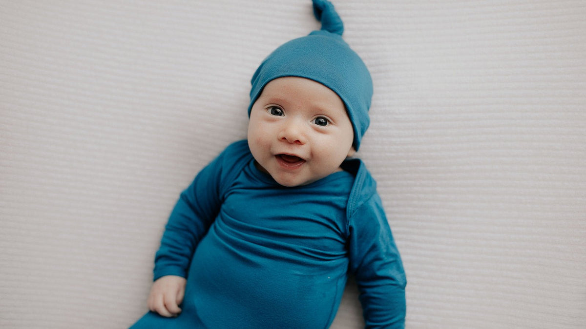 Smiling baby boy in a blue knotted gown and matching hat lying on a white blanket