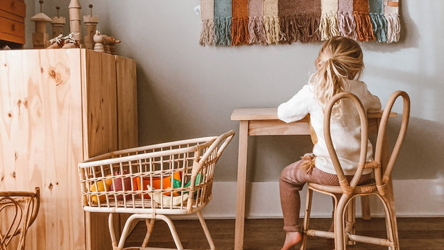Preschooler drawing at a child-size wooden table in a calm playroom with a rattan doll crib and wooden toys