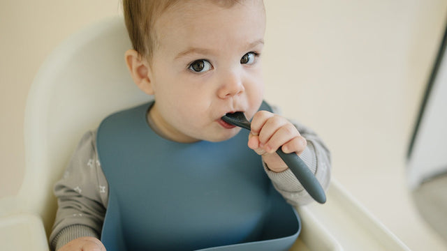 Baby sitting upright in a high chair with a silicone bib, holding a soft-tipped spoon in his mouth