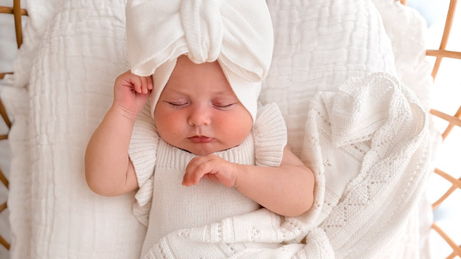 Newborn wearing a white bow turban asleep on her back in a rattan bassinet, tucked under a white pointelle knit blanket