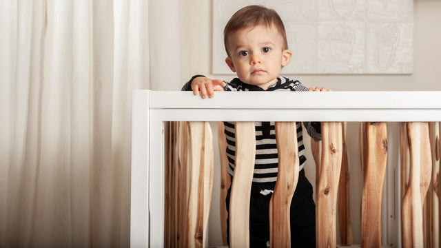 Baby in a striped hoodie standing up in a white and natural-wood crib in a bright nursery