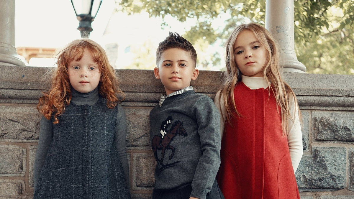 Three children in Cottier kids clothing: a charcoal wool jumper, an equestrian sweater and a crimson pinafore dress, standing by a stone wall