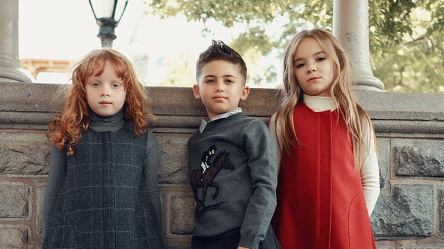 Three children in Cottier kids clothing: a charcoal wool jumper, an equestrian sweater and a crimson pinafore dress, standing by a stone wall