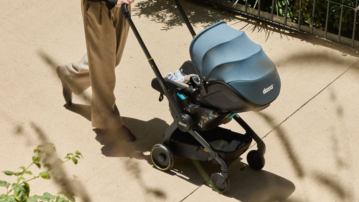 Parent pushing a baby along a sunlit path in a slate-blue Doona car seat stroller, wheels rolled down and canopy up