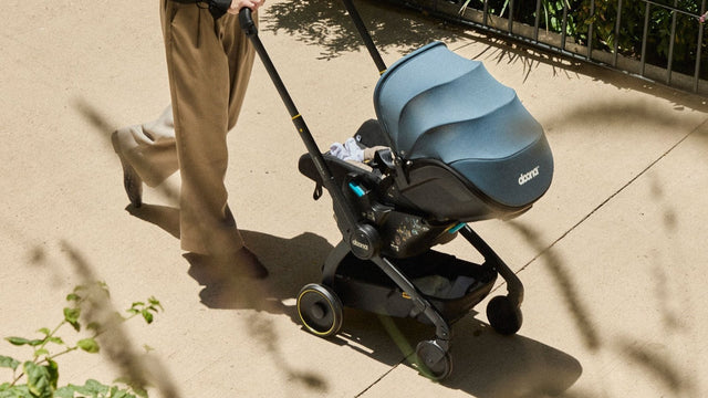 Parent pushing a baby along a sunlit path in a slate-blue Doona car seat stroller, wheels rolled down and canopy up