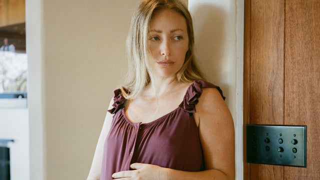 Pregnant woman in a plum labour-and-delivery gown standing in a sunlit doorway at home, one hand resting on her bump