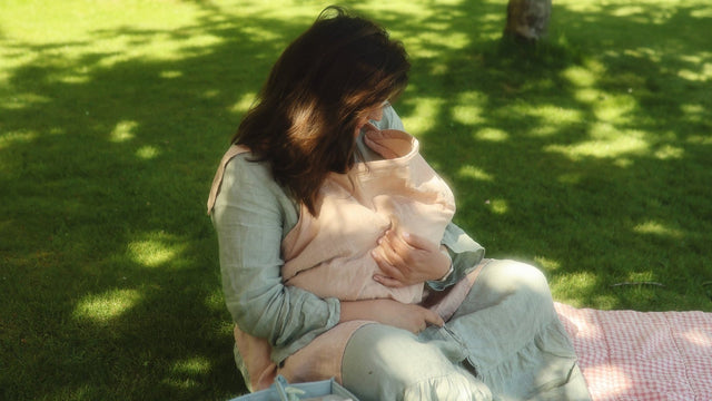 Mother breastfeeding her baby under a muslin nursing cover on a picnic blanket in a sunlit garden, nursing caddy beside her