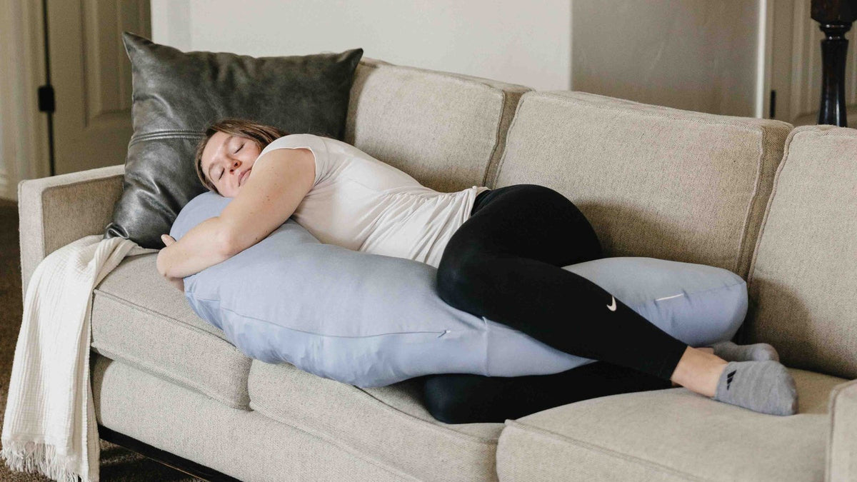 Pregnant woman napping on a living-room sofa, curled around a body pillow during first-trimester fatigue