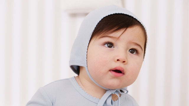 Baby in a pale blue pointelle bonnet and matching top looking up, in a bright nursery