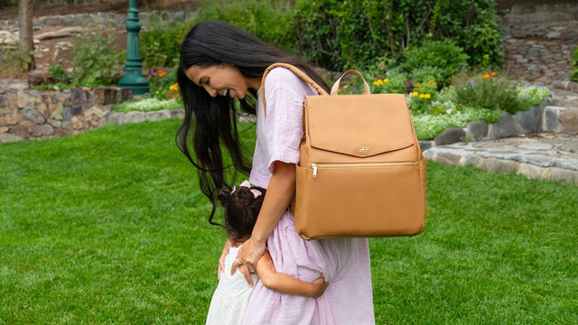 Mother wearing a butterscotch Freshly Picked Classic Diaper Bag II as a backpack while hugging her toddler on a garden lawn