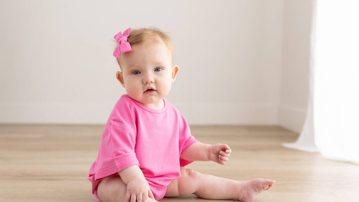 One-year-old girl in a bright pink romper and matching hair bow sitting on a wood floor