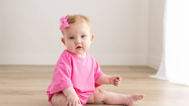 One-year-old girl in a bright pink romper and matching hair bow sitting on a wood floor