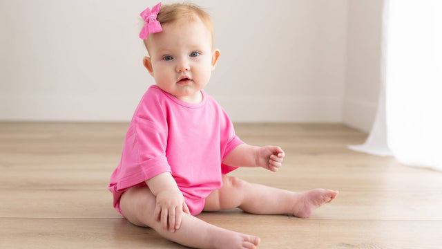 Baby girl in a bright pink romper and bow sitting on a wood floor