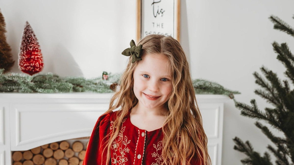 Girl in a red embroidered corduroy Christmas dress beside a holiday mantel and tree