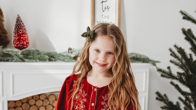 Girl in a red embroidered corduroy Christmas dress beside a holiday mantel and tree