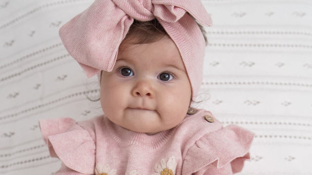 Baby girl in a dusty-pink knit romper and oversized bow headwrap sitting on a white quilted bed