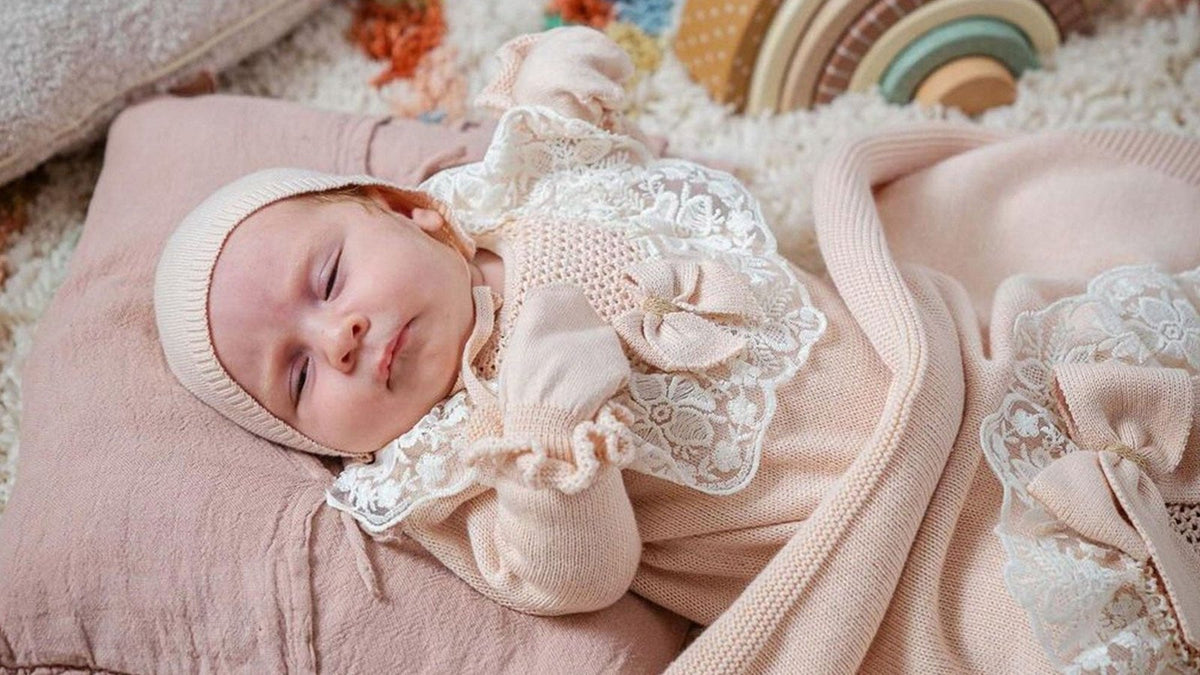 Newborn in a knit bonnet and lace-trimmed outfit resting on a pink linen cushion under a soft knit blanket