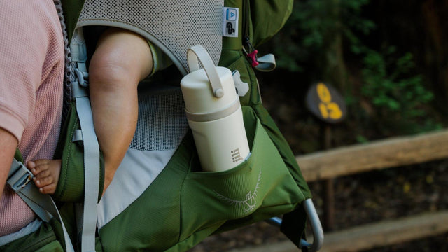 Jili all-in-one insulated baby bottle tucked in a hiking carrier pocket next to a baby's foot on a trail
