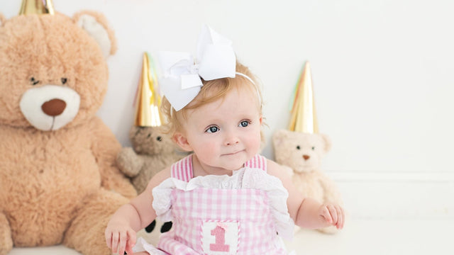 Baby in a pink gingham number-one birthday outfit with party hats and teddy bears