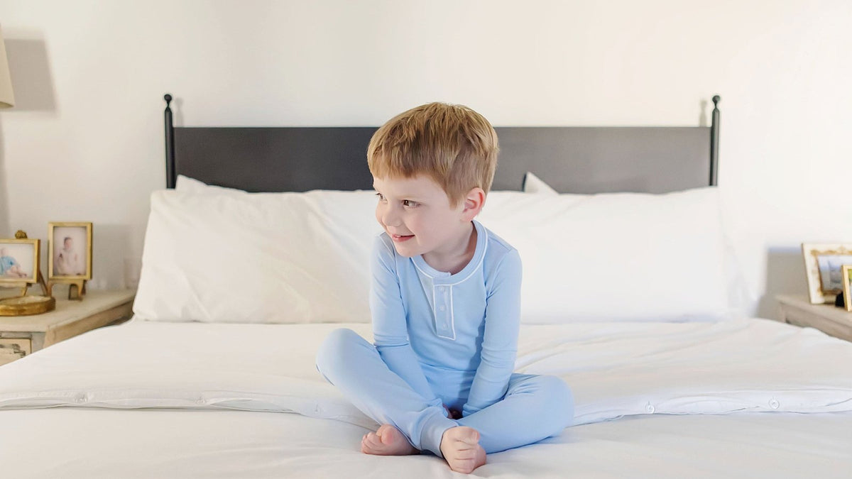 Smiling boy sitting cross-legged on a white bed in light blue Lila + Hayes pima cotton pajamas