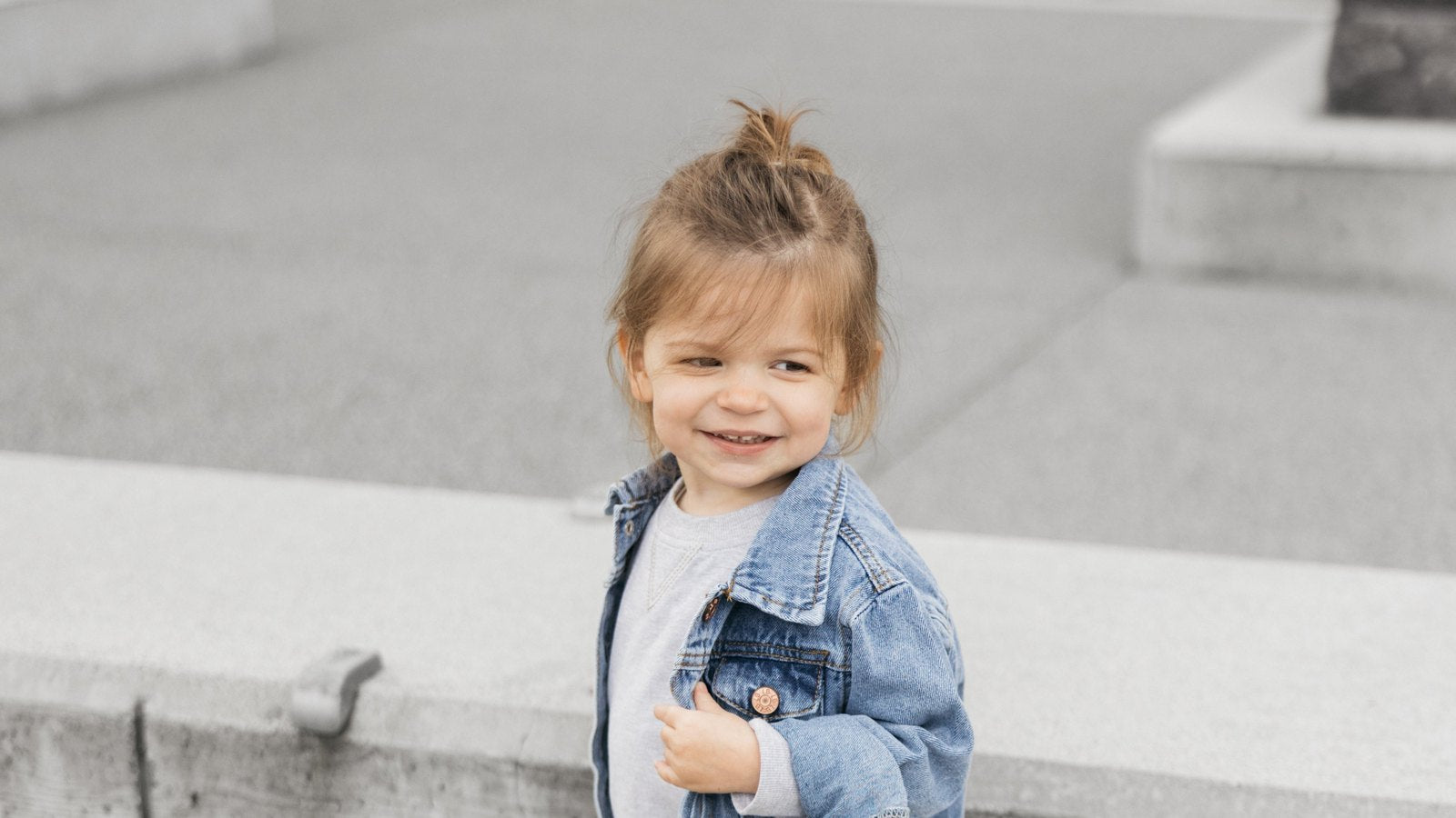 Smiling toddler girl wearing a Little Bipsy denim jacket over a grey sweatshirt and joggers outdoors