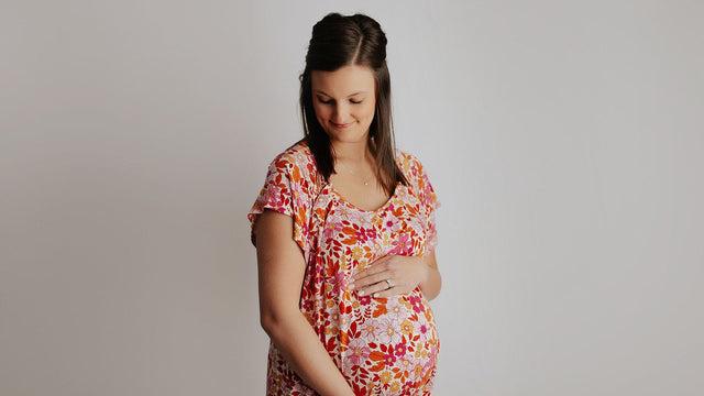 Pregnant woman in a pink and orange floral maternity dress smiling down at her bump, both hands cradling her belly