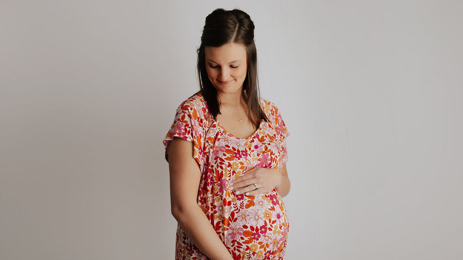 Pregnant woman in a pink and orange floral maternity dress smiling down at her bump, both hands cradling her belly