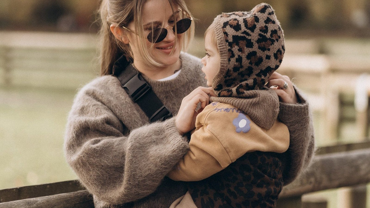 Mother holding her baby close outdoors, a carrier strap across her sweater and the baby in a leopard-print knit bonnet