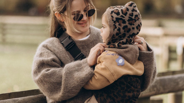 Mother holding her baby close outdoors, a carrier strap across her sweater and the baby in a leopard-print knit bonnet