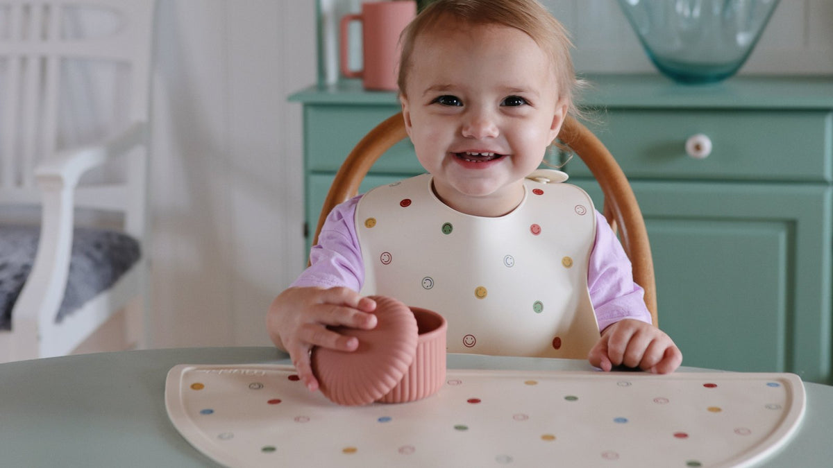 Smiling toddler wearing a Mushie silicone bib at the table with a matching Mushie snack cup and placemat