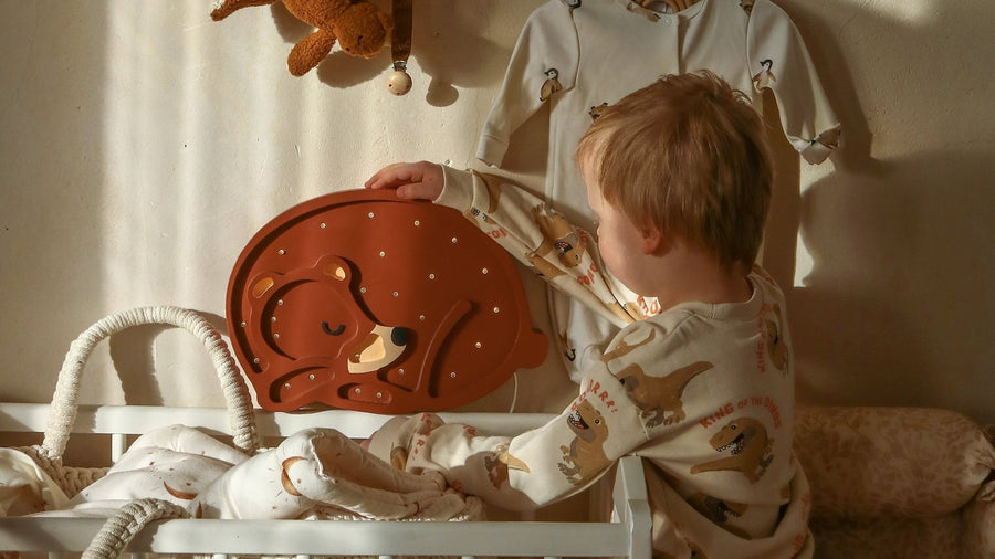 Toddler in pajamas standing at a white crib in a sunlit nursery, reaching for a wooden sleeping-bear night light on the crib rail