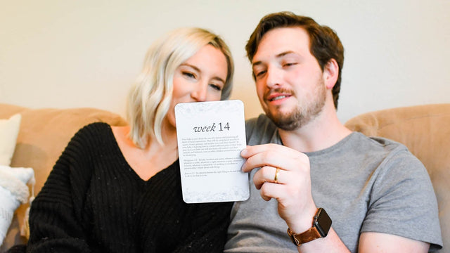 Smiling couple on the sofa holding up a week-14 pregnancy milestone card