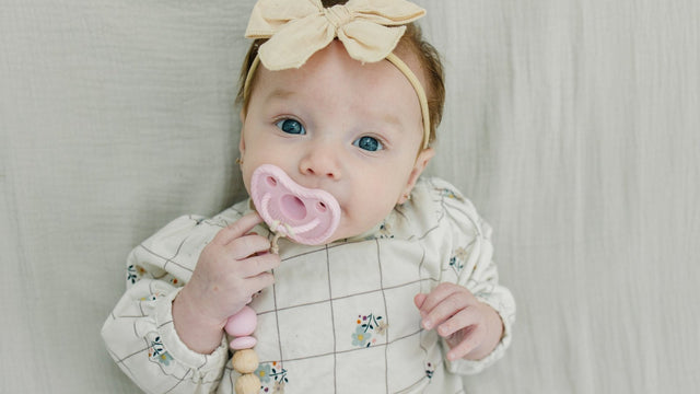 Baby girl with a bow headband soothing herself with a pink round silicone pacifier