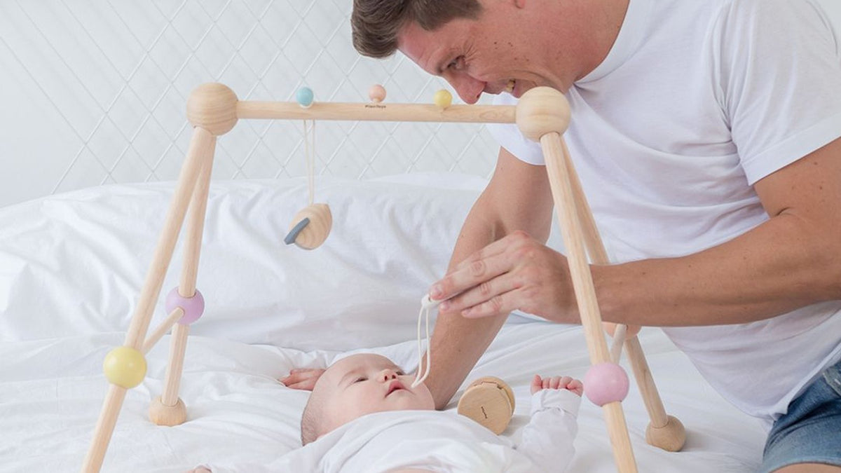 Dad playing with baby under a PLAN Toys wooden play gym made from sustainable rubberwood