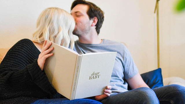 Couple sharing a kiss on the sofa at home, half-hidden behind a keepsake baby journal