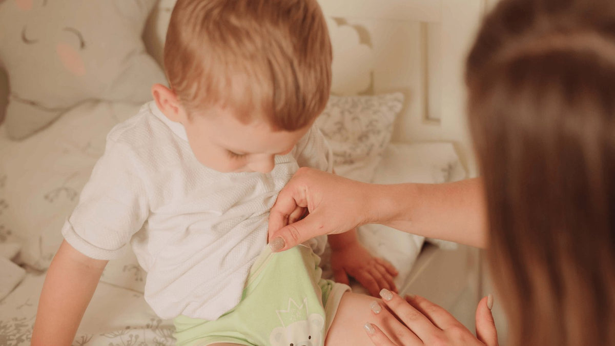 Parent helping a toddler pull up soft green training pants on the bed during potty training