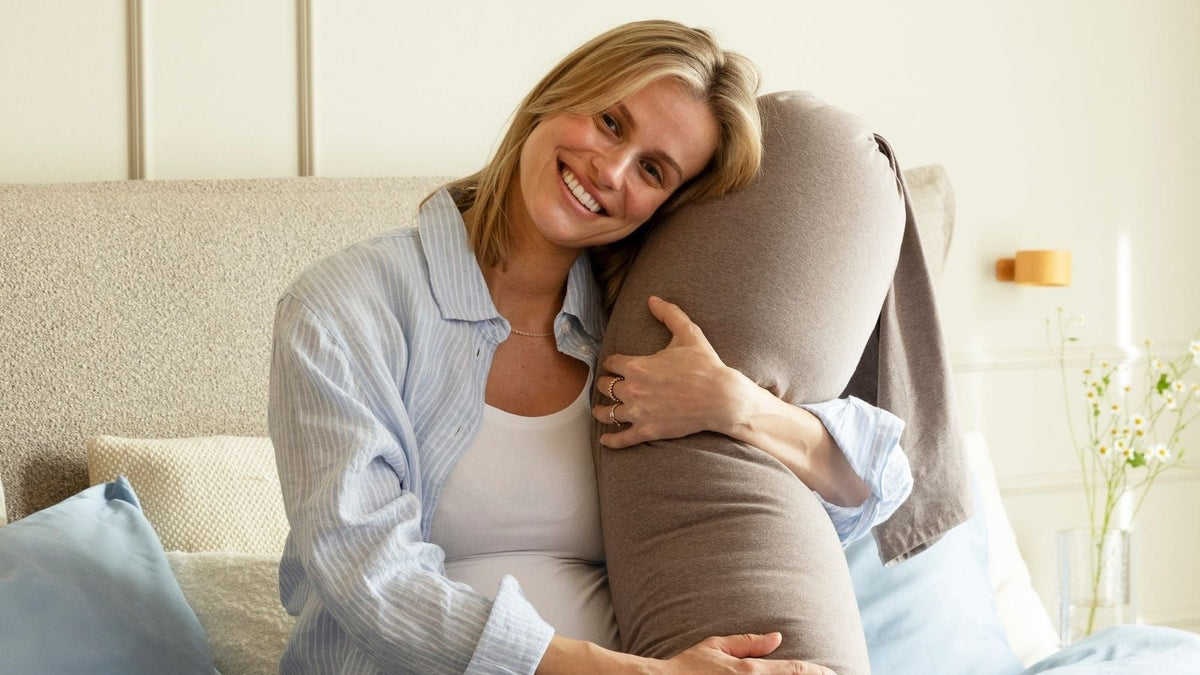 Pregnant woman in pyjamas sitting up in bed, resting her head against a full-length pregnancy support pillow