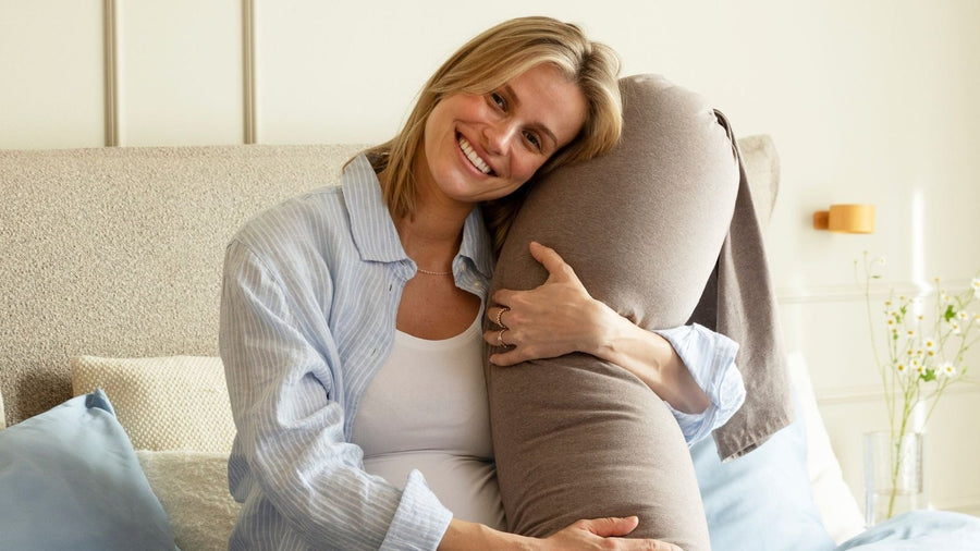 Pregnant woman in pyjamas sitting up in bed, resting her head against a full-length pregnancy support pillow