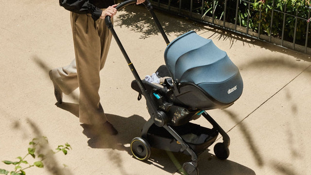Parent walking a baby along a sunny path in a Doona infant car seat rolled out in stroller mode, canopy up