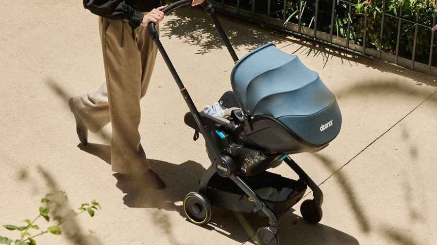 Parent walking a baby along a sunny path in a Doona infant car seat rolled out in stroller mode, canopy up
