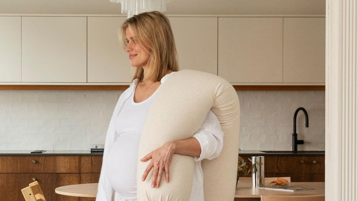 Pregnant woman with a second-trimester bump standing in a bright kitchen, holding a full-length pregnancy support pillow