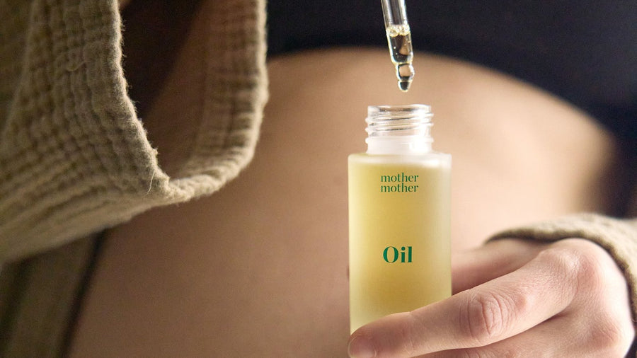 Close-up of a bare pregnancy bump with a hand holding a dropper bottle of nourishing belly oil