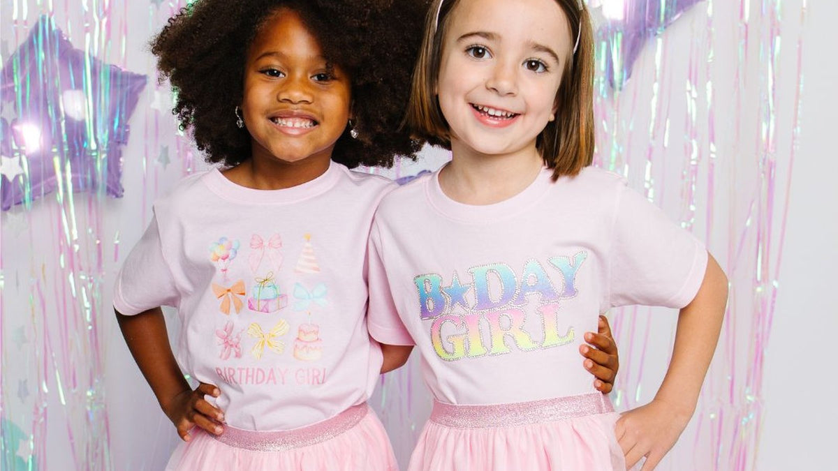 Two smiling girls in Sweet Wink birthday girl t-shirts and pastel confetti tutus at a balloon-filled party