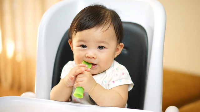 Baby sitting in a high chair chewing on a soft green silicone training spoon