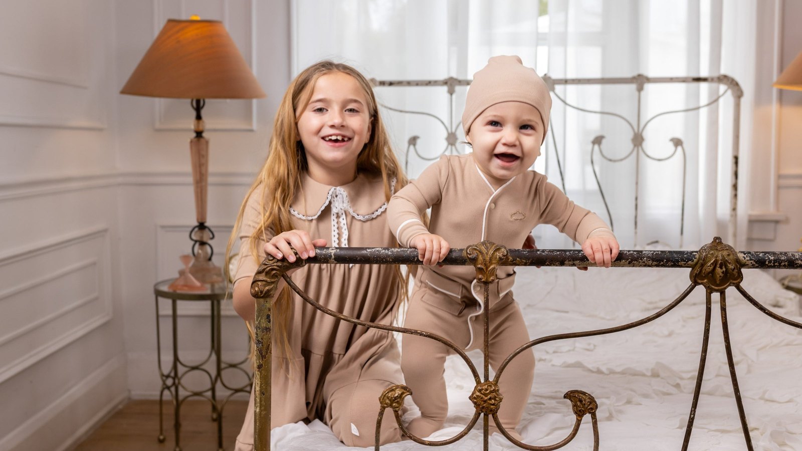 Laughing baby in a cocoa ribbed footie pajama and matching beanie standing at an iron bed frame beside a big sister in a bedroom