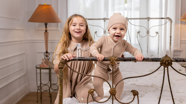Laughing baby in a cocoa ribbed footie pajama and matching beanie standing at an iron bed frame beside a big sister in a bedroom