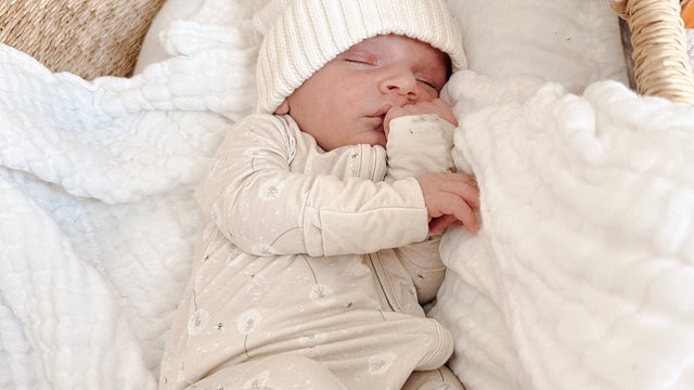 Newborn asleep in a woven moses basket wearing a cream bamboo zip-up footie pajama with a dandelion print and a knit pom-pom hat